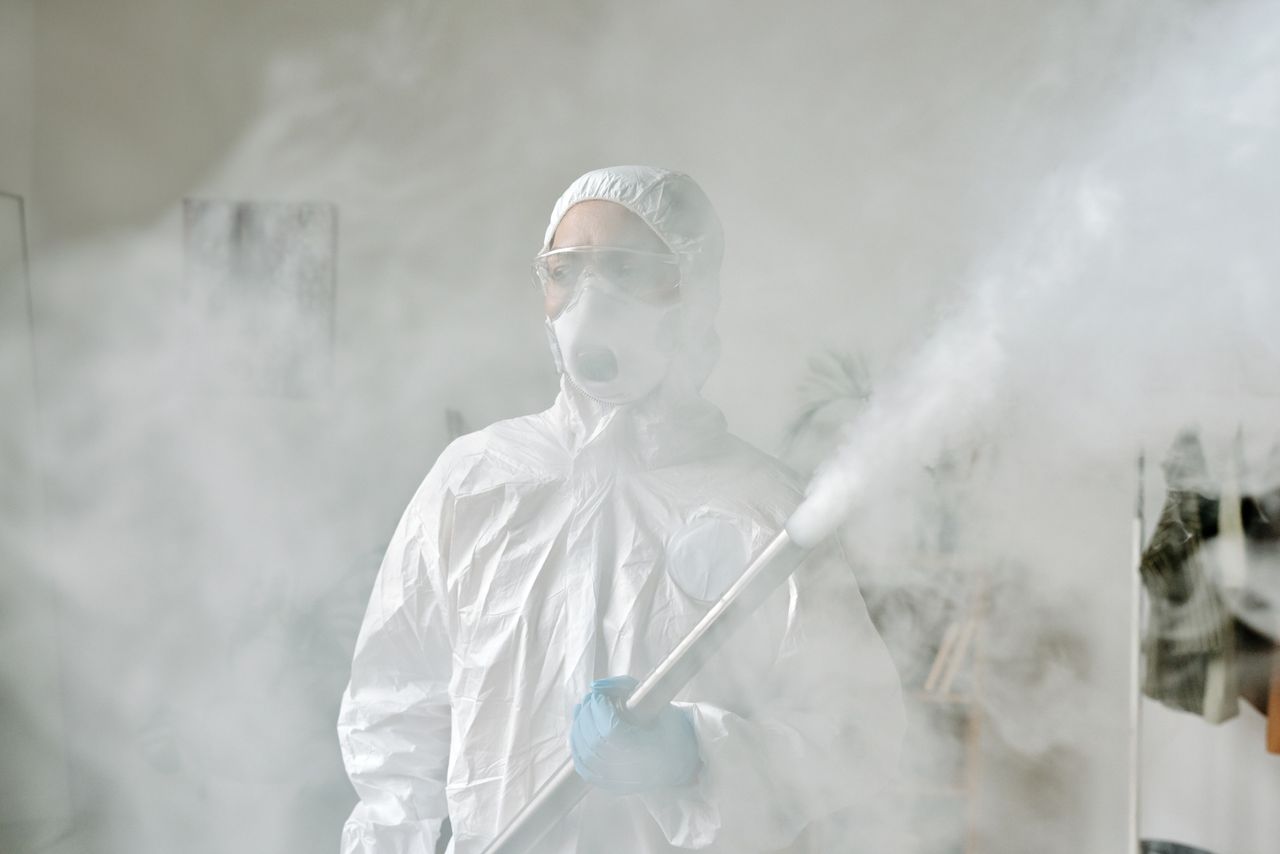 brand-02 Person in protective gear fumigating an area indoors amid smoke for disinfection.